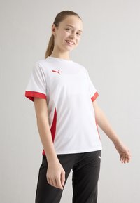 Valge spordisärk punaste detailidega, millel on lühikesed varrukad, hingavad võrkdetailid külgedel ja logo vasakul rinnal, kombineeritud mustade pükstega.