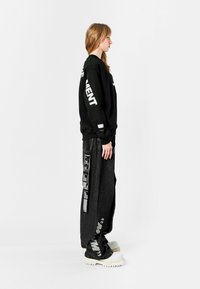 Sudadera negra con texto blanco en las mangas, combinada con pantalones grises anchos que presentan gráficos llamativos y un acabado texturizado. Zapatillas blancas voluminosas.