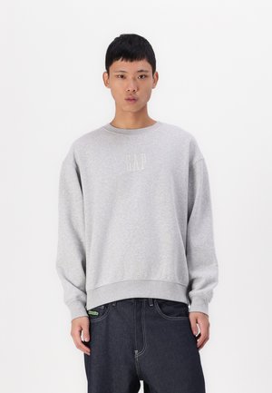 Sweatshirt gris avec un col rond côtelé, arborant un petit logo "GAP" en couleur claire. Associé à un jean bleu foncé.