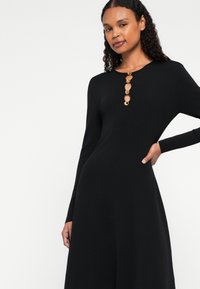 Čierne šaty s dlhými rukávmi a vysokým golierom, s radom piatich okrúhlych zlatých gombíkov, hladkou textúrou a priliehavým strihom.