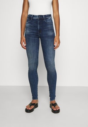 Jean skinny en denim bleu foncé avec une texture lisse, doté d'une taille mi-haute et d'un design classique à cinq poches, associé à des sandales noires plates.