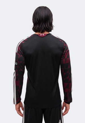 Persona con el cabello oscuro hasta los hombros que lleva una camiseta deportiva negra de manga larga con mangas estampadas en granate y negro y rayas blancas.