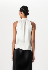Blouse sans manches, gris clair en tissu lisse avec un col montant. Présente des détails plissés et un ourlet arrondi, associée à une jupe plissée noire.