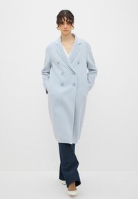  Lichtblauwe dubbele rij knoopjas met ronde knopen, gemaakt van zachte stof. Heeft een iets oversized pasvorm en zijzakken, gecombineerd met donkere jeans.