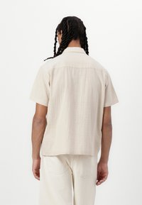 Camisa de manga corta en un tejido de color beige claro, con corte recto y detalle en el pezón trasero. Llevada con pantalones de color claro a juego.