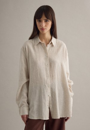 Jonge vrouw met lang donker haar, gekleed in een loszittend beige overhemd met knopen en een bruine broek, tegen een effen lichtgrijze achtergrond.