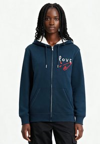 Haut à capuche zippé bleu marine avec doublure en polaire blanche, arborant un motif avant avec le mot "love" en rouge et blanc. Comprend des poches kangourou.