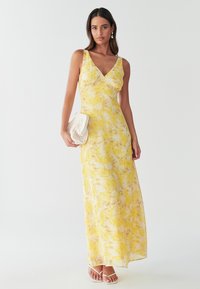 Yellow, floral-patterned maxi dress with a deep V-neck, thin straps, and a flowy silhouette. Paired with a cream-colored clutch and white sandals.