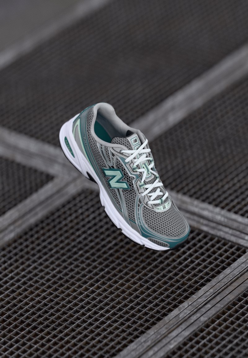 Baskets de course New Balance grises et teal avec des lacets blancs posées sur une surface en grille métallique.