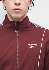 Bordowa kurtka na zamek z wysokim kołnierzem, białymi lamówkami i logo Reebok. Gładki materiał o dopasowanym kroju z kontrastowymi ciemnymi panelami na ramionach.