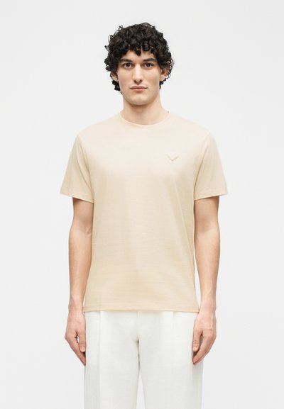 Άνδρας με σγουρά σκουρόχρωμα μαλλιά, φορώντας απλό μπεζ κοντομάνικο μπλουζάκι και λευκό παντελόνι, στέκεται με το σώμα στραμμένο προς τα εμπρός σε λευκό φόντο.