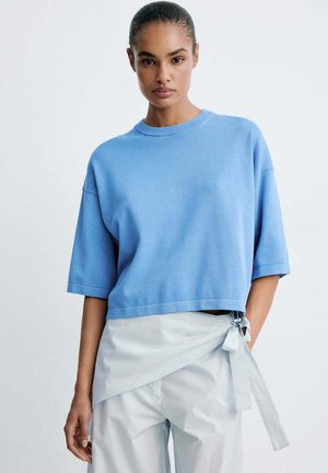 Femme aux cheveux tirés en arrière portant un pull bleu clair à manches courtes et un pantalon blanc style portefeuille noué sur le côté.