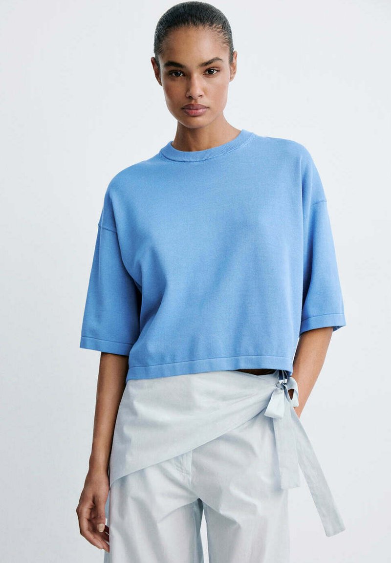 Femme aux cheveux tirés en arrière portant un pull bleu clair à manches courtes et un pantalon blanc style portefeuille noué sur le côté.