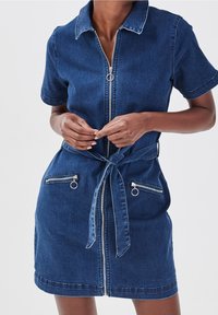 Robe en jean bleu foncé, avec une fermeture éclair frontale, des manches courtes et deux poches zippées avec anneaux de tirage. Ceinture nouée fuselée à la taille.