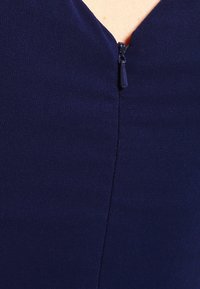 Marinblå klänning med V-ringning, med en centrerad dragkedja. Tyget har en slät struktur med en subtil lyster.