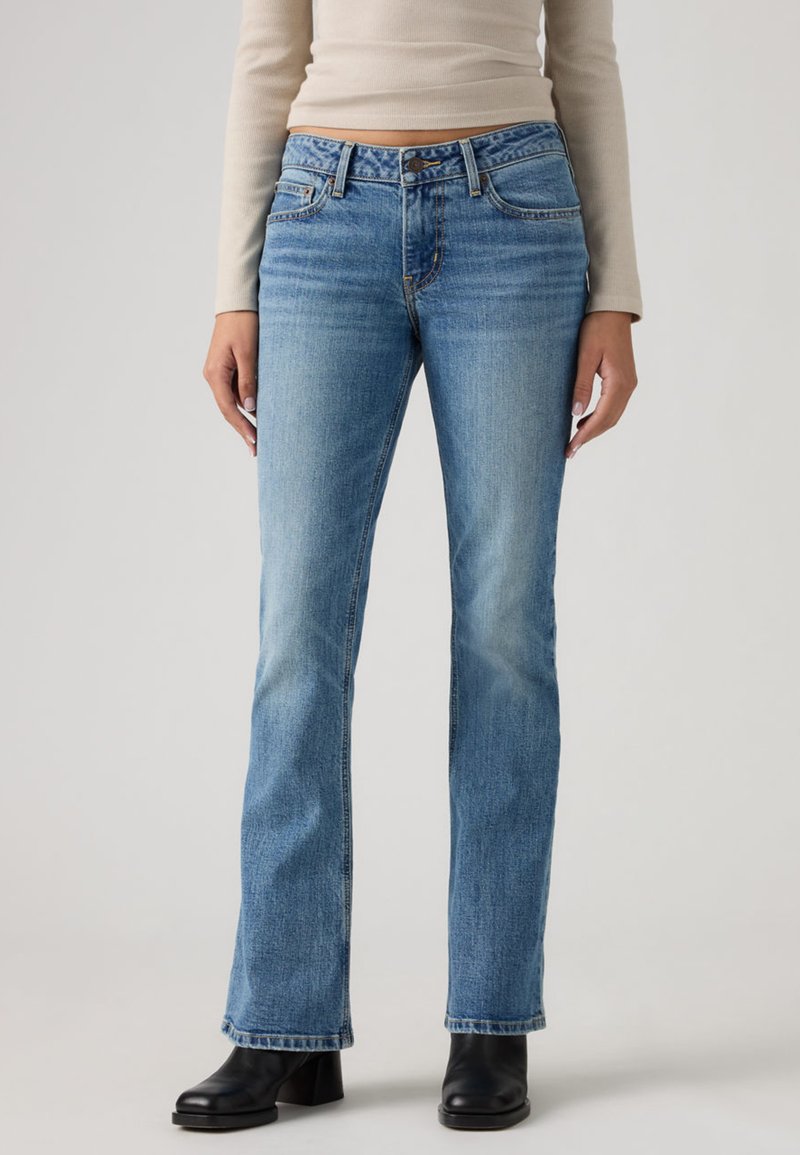 Άτομο που φοράει ανοιχτό μπλε τζιν παντελόνι με ίσια μπατζάκια, μαύρα μποτάκια μέχρι τον αστράγαλο και μπεζ μακρυμάνικη μπλούζα, στέκεται μπροστά σε απλό φόντο.