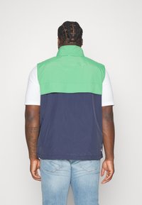 Farbblockierte Weste mit einem oberen Teil in Grün und einem unteren Teil in Marineblau, aus leichtem Material gefertigt; mit einem hohen Kragen und ohne sichtbare Beschläge.