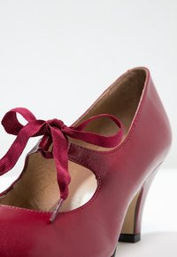 Chaussure à talon haut en cuir rouge avec un nœud à l'avant, design découpé et texture lisse. Le talon est noir contrastant.