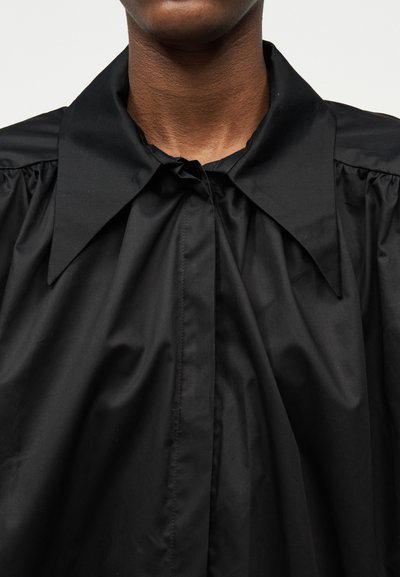 Personne à la peau foncée portant une chemise noire avec un col surdimensionné et pointu ainsi que des épaules froncées.