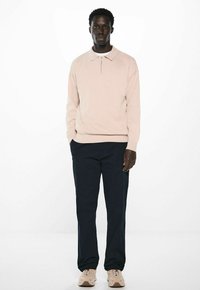 Helles rosa Zip-Up Sweatshirt mit langen Ärmeln, getragen mit marineblauen Hosen und beigen Sneakers auf einem schlichten weißen Hintergrund.