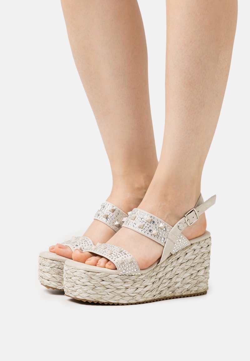 Pieds portant des sandales compensées à plateforme tissée beige avec des brides cloutées et une fermeture à boucle sur fond blanc.