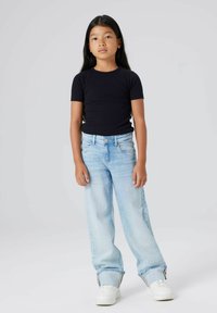 Ung jente står, iført en svart T-skjorte med korte ermer, lyseblå vide jeans med oppbrett, og hvite sneakers foran en ensfarget grå bakgrunn.