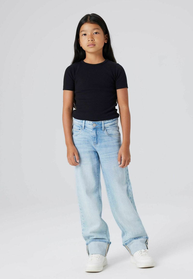 Ung jente står, iført en svart T-skjorte med korte ermer, lyseblå vide jeans med oppbrett, og hvite sneakers foran en ensfarget grå bakgrunn.