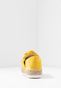 Espadrille amarillo de ante con media suela de yute trenzado y suela exterior de goma blanca estriada, mostrada desde atrás sobre una superficie blanca y un fondo de esquina de pared.