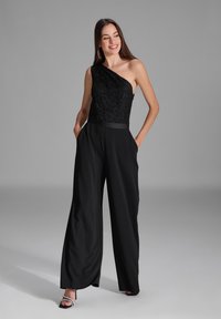 Schwarzer Jumpsuit mit einem Schulterdesign, das ein strukturiertes Spitzenoberteil und weite Hosenbeine aufweist. Enthält Taschen und einen glatten Taillenbund.