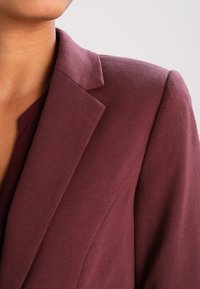 Gros plan d'une personne portant un blazer bordeaux sur mesure à revers à crans, avec un haut assorti, sur un fond clair.