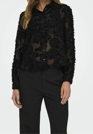 Woman wearing semi-sheer black button-up blouse with textured floral patterns paired with high-waist black trousers, standing against white background.