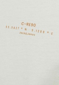 White cotton fabric with printed text in light brown. Text includes coordinates and brand name "Jack & Jones" with a clean, minimalist design.