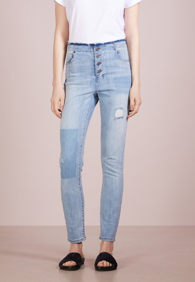 Personne portant un jean skinny taille haute bleu clair avec fermeture à boutons, des patchs usés, un t-shirt blanc et des sandales noires à bout ouvert.