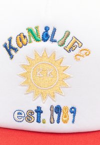 Valge müts, millel on värvikas tikand tekst "KANILIFE" ja "est. 1989," koos kollase päikese graafika ja punaste aktsentidega.