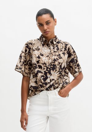 Vrouw die een bruin-beige gebloemde korte mouwen blouse met knopen draagt, ingestopt in een wit broek met hoge taille, staat met één hand in de zak.