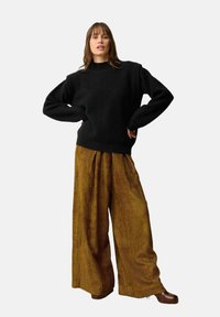 Czarny sweter o teksturowanej fakturze z długimi rękawami w połączeniu z szerokimi, prążkowanymi spodniami z brązowego kordurowego materiału. Modelka stoi z rękami na biodrach na białym tle.