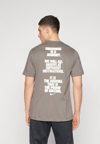 Camiseta gris de manga corta con gran texto blanco en la parte posterior. El texto incluye frases motivacionales y un logo de Nike en la parte inferior.