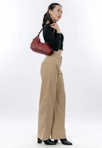 Rode leren schoudertas met een gestructureerd oppervlak, gouden hardware en een compact formaat. Model draagt een zwarte crop top en beige wijde broek.