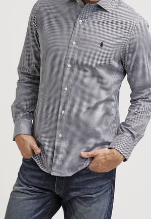 Homme portant une chemise à manches longues à petits carreaux noir et blanc avec un jean bleu foncé, mains dans les poches.