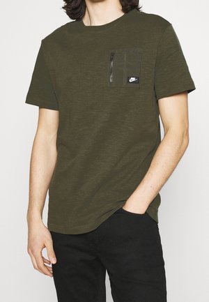 Homme portant un t-shirt à manches courtes vert olive avec une poche chest zippée et le logo Nike, associé à un pantalon noir, main dans la poche.