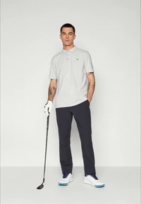 Grå polo-skjorte med tre knapper, mørke bukser, hvite golfesko med blå detaljer og en golfkølle, stående mot en ensfarget bakgrunn.