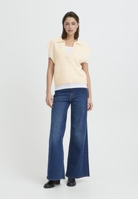 Gilet en tricot pêche clair avec un col en V et des manches courtes, superposé sur une chemise blanche, associé à un jean en denim bleu foncé à jambe large et des bottes noires.