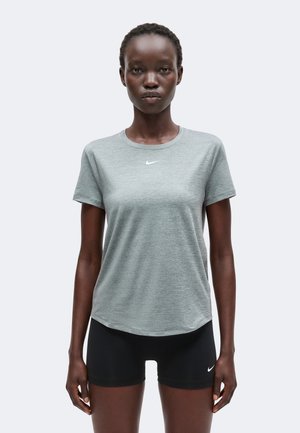 Kobieta stoi twarzą do przodu, ubrana w szarą koszulkę Nike i czarne krótkie spodenki Nike na jednolitym jasnym tle.