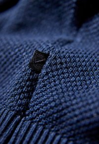Tessuto lavorato a maglia blu navy con un motivo testurizzato e sezioni a coste prominenti, completato da un piccolo etichetta nera con un design geometrico.