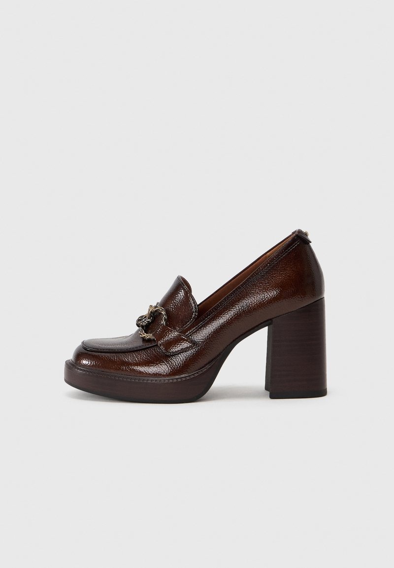 Mocassin en cuir marron foncé avec talon haut épais et détail décoratif en chaîne métallique sur le devant, présenté de profil sur un fond uni.
