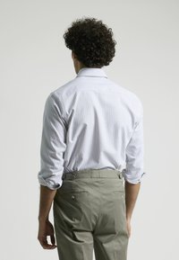 Camisa a rayas blanca y azul con mangas largas, metida dentro de pantalones verdes oliva con correas laterales ajustables y bolsillos traseros.