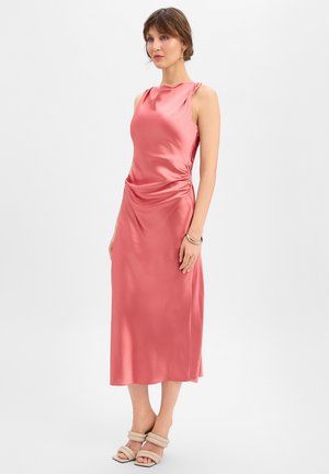 Frau trägt ein ärmelloses, glänzendes, korallrosa Midikleid mit gerafften Seiten und beigefarbenen, doppelt gespassten, offenen Schuhen mit Absatz, steht vor einem einfarbigen Hintergrund.