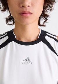 Witte sportieve tanktop met zwarte accenten en drie zwarte strepen op de schouders. Beschikt over een zilveren Adidas-logo op de borst.