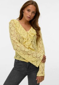 Donna che indossa un top lungo a maniche con pizzo floreale giallo e rouches, abbinato a jeans grigi scuri, in posa con le braccia incrociate su sfondo bianco.