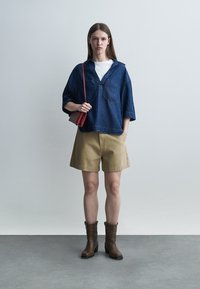 Žena oblečená v rifľovej bunde cez biele tričko, béžových šortkách, hnedých čižmách a držiaca malú červenú kabelku stojaca pred jednofarebným pozadím.